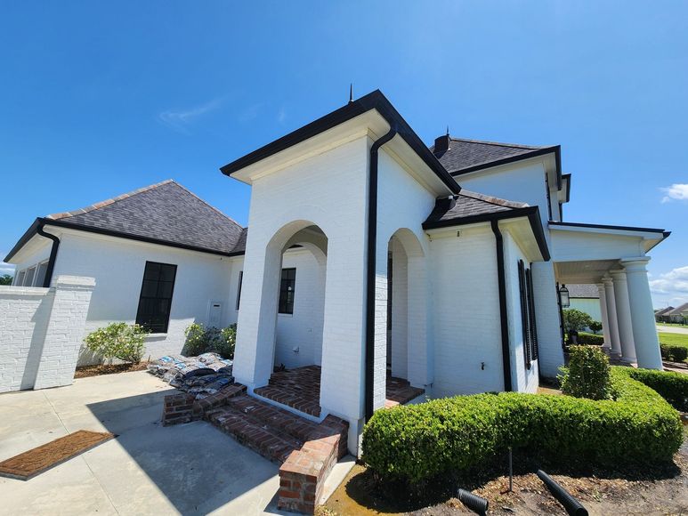 White stucco house with black trim and roof. Green hedges border the front yard, under a blue sky.