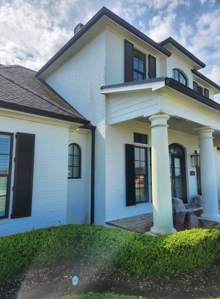 White brick house with black shutters and roof; a front porch with large white columns. Green bushes in front.