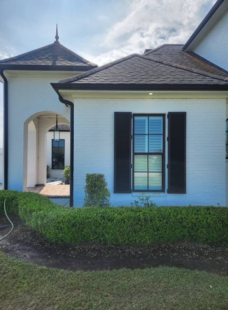 White brick house with dark shutters and a manicured hedge under a bright sky. A covered entryway is visible.