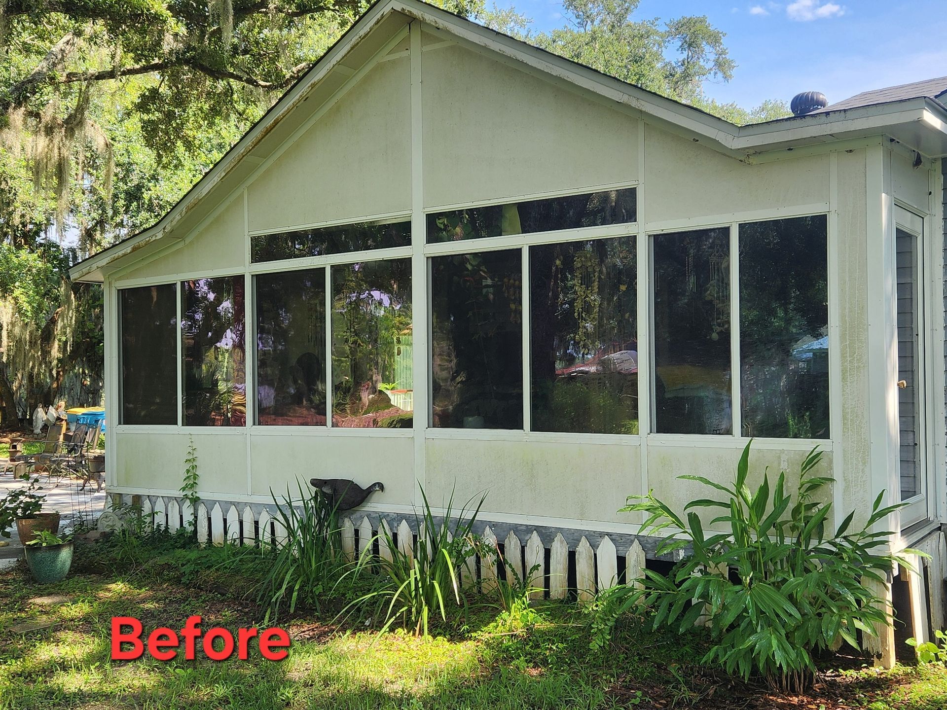 A before shot of a screened-in porch with white siding and large windows. The yard in front has overgrown plants.