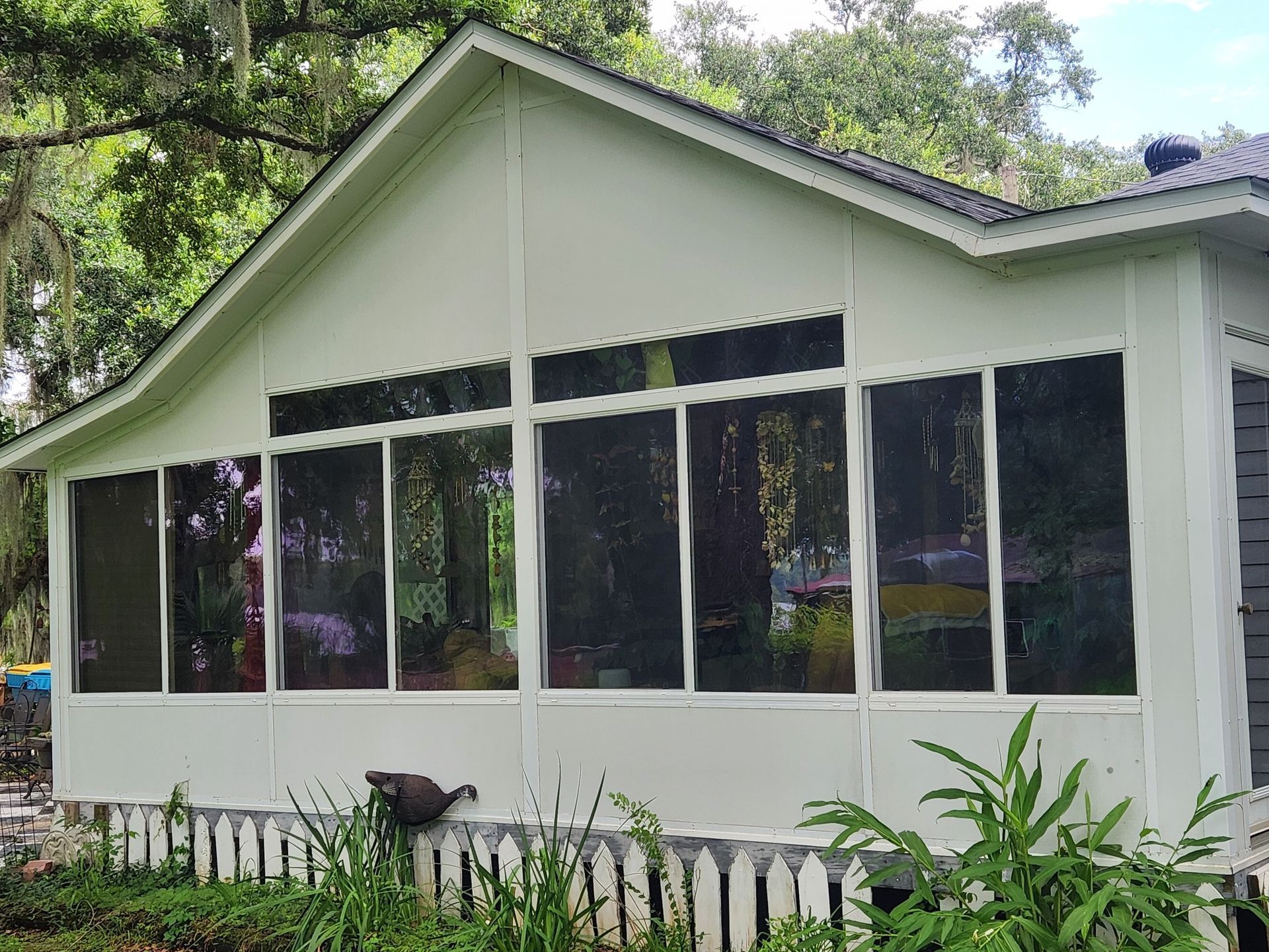 White screened-in porch with large windows, overlooking a yard with plants and a white picket fence.