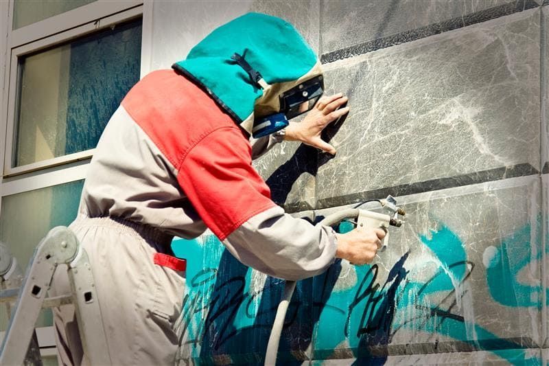Person wearing protective gear sprays graffiti on a building, standing on a ladder. They wear a red and grey suit and a green hood.