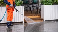 A worker in orange coveralls power washes a wet, dark gray sidewalk outside a cafe.