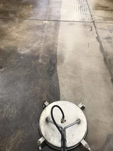A circular power washer cleaning a concrete surface, leaving a clean path behind. The unwashed area is dark and dirty.