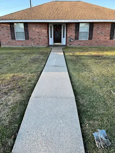 A concrete walkway leads to a brick house with a brown roof. The lawn is green and brown, and work gloves rest on the side.