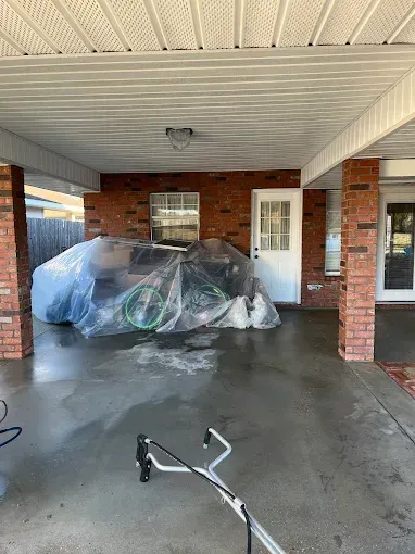 A carport with wet concrete floor. A car covered in plastic, and a pressure washer are visible. Brick pillars support the roof.