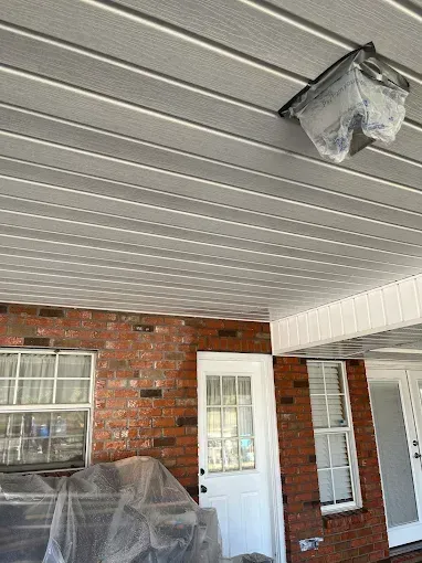 View from below of a porch ceiling with gray paneling, a brick wall, windows, and a light fixture covered in plastic.