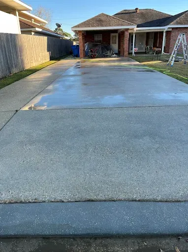 A wet, freshly-cleaned concrete driveway leads to a red brick house.  A wooden fence and grass are visible on the left.