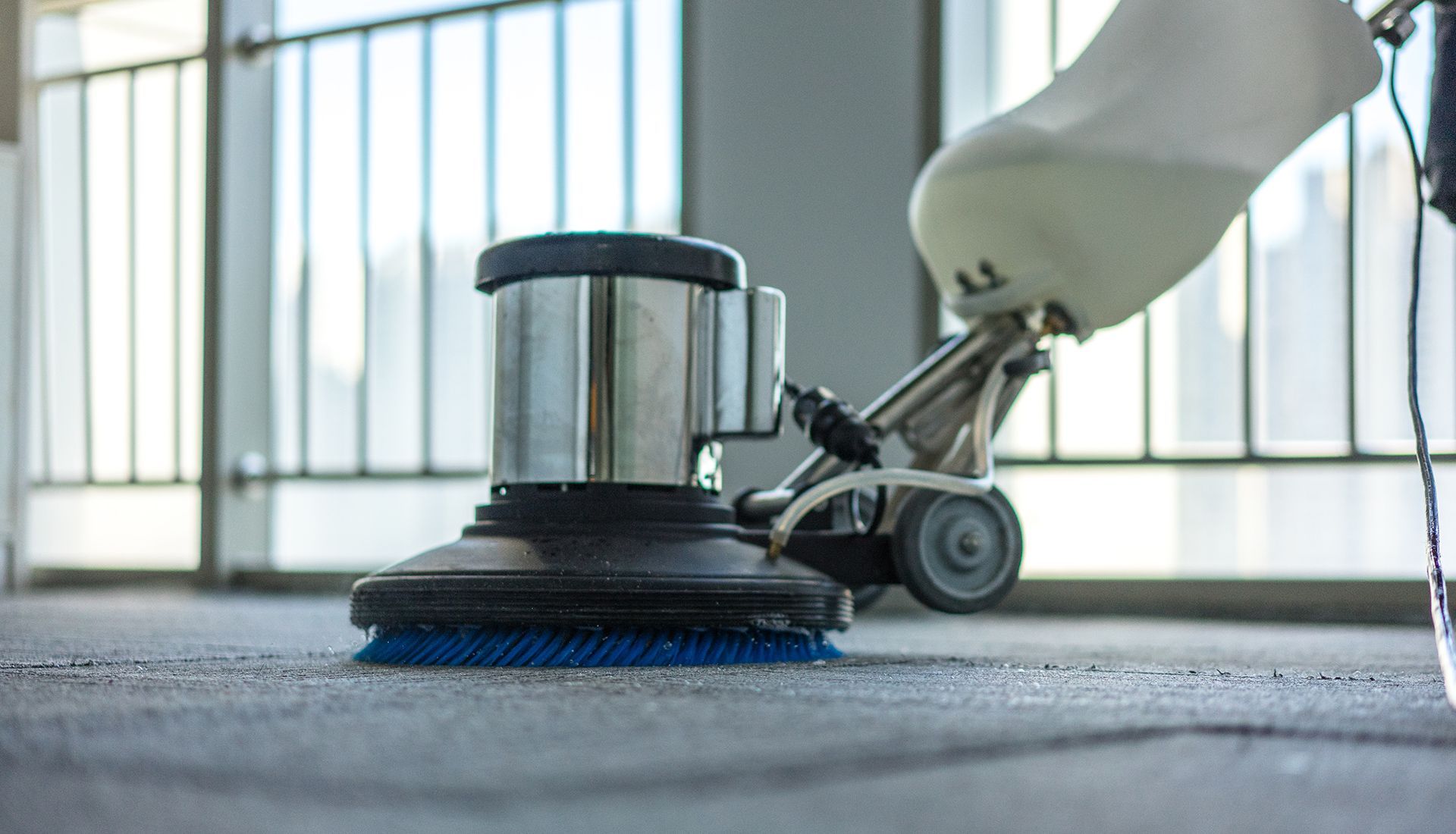 Floor cleaning machine scrubbing a gray carpet in an office with large windows.