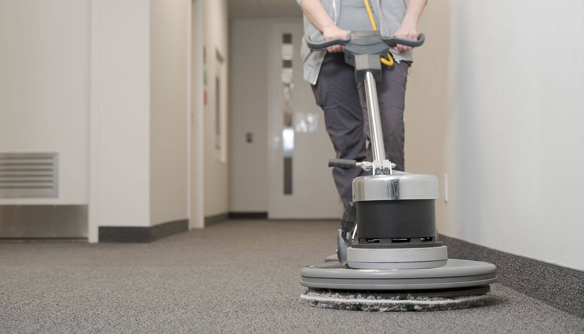 Person operating a floor cleaning machine on a gray carpeted hallway.