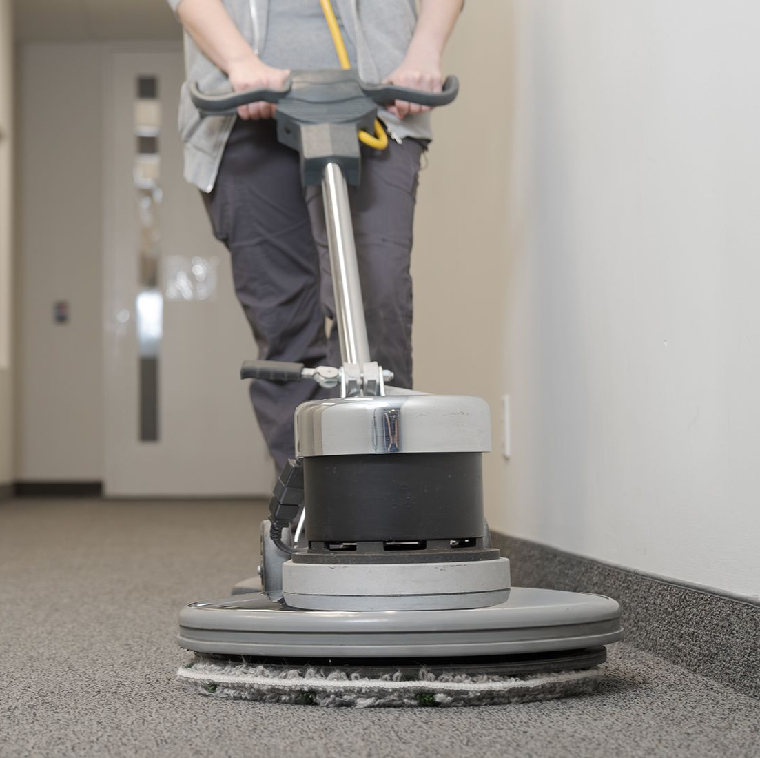 Person using floor buffer on carpeted hallway floor.