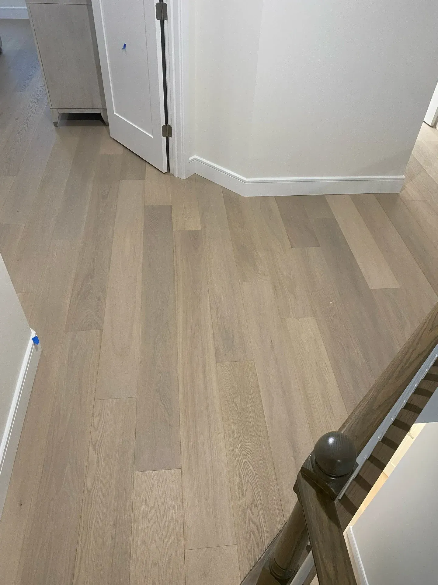 Light wood floor in a hallway with white walls, a doorway, and a stair railing visible.
