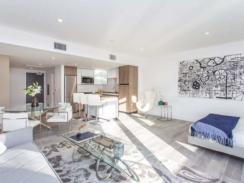 Modern open-concept apartment with kitchen, living area, and bed. White and gray decor, bright sunlight.