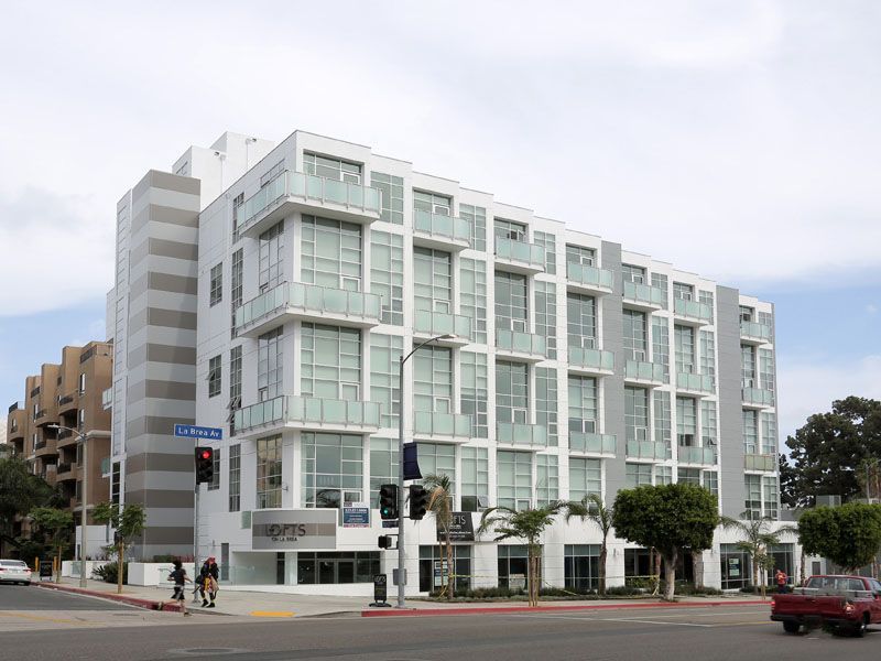 Modern, multi-story white building with glass balconies, at a street intersection. Two cyclists.