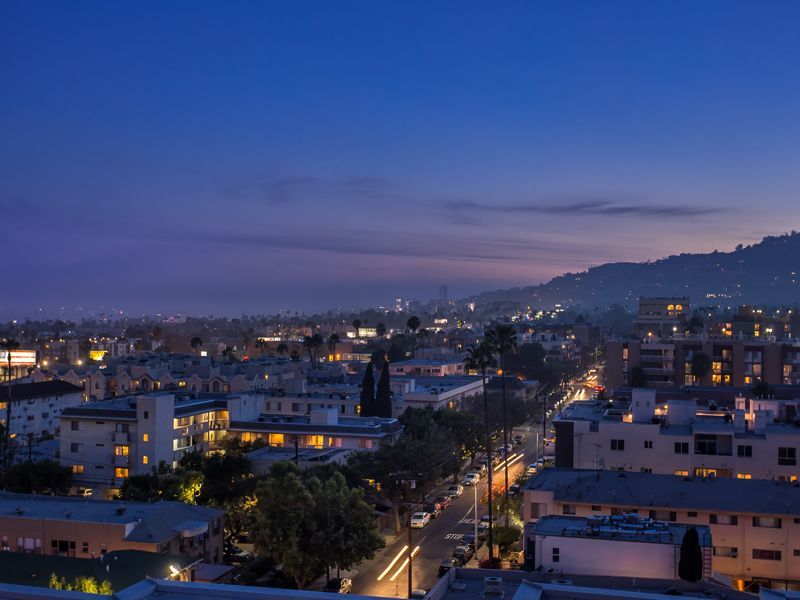 City skyline at dusk with lights, buildings, and a road. Hilly area in the background under a blue sky.