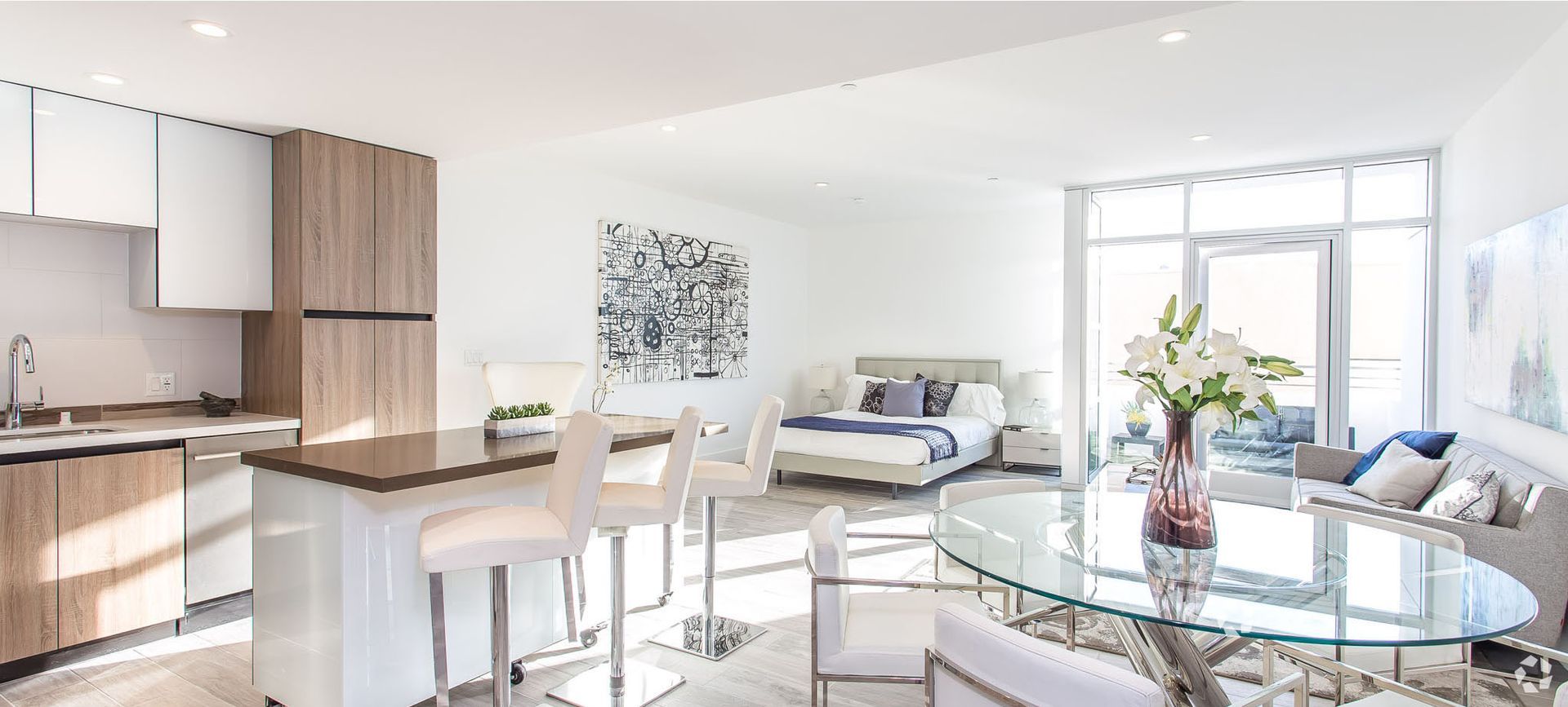 Modern open-concept apartment with kitchen, dining area, and bedroom. Bright white walls, wood cabinets, glass table, and large windows.
