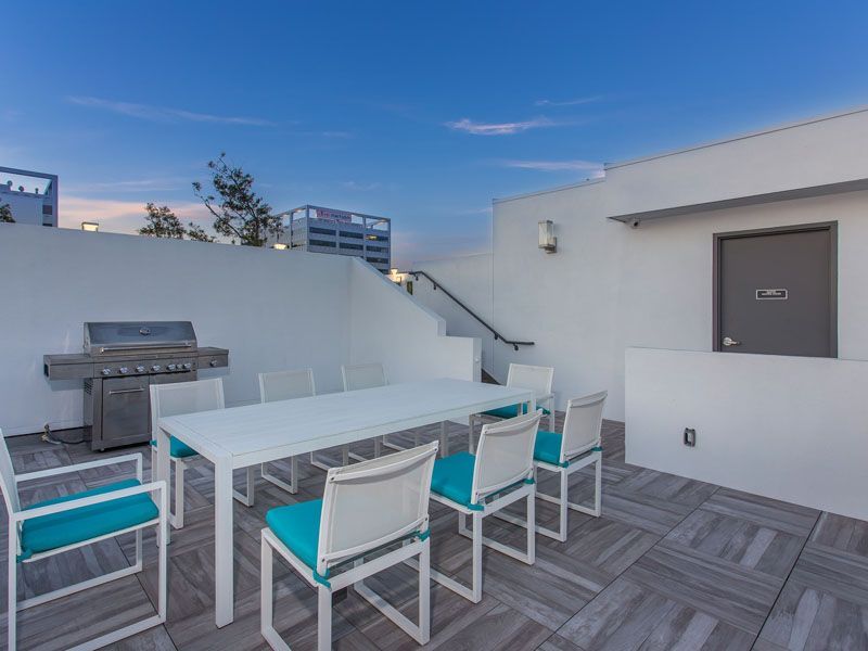 Rooftop patio with white table, turquoise chairs, grill, and stairs, against a blue sky.