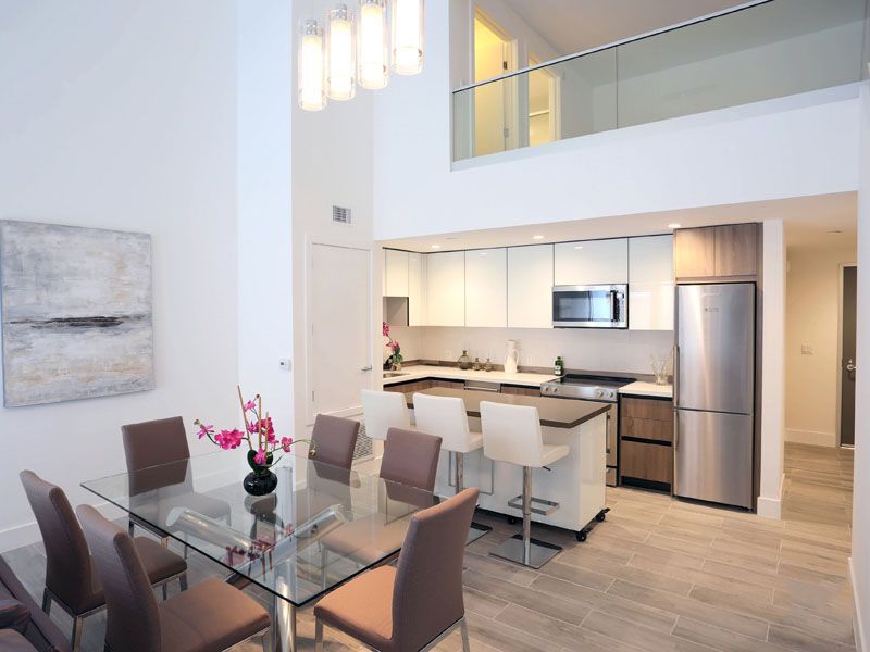 Dining room with glass table, adjacent to an open kitchen with white cabinets and stainless steel appliances.