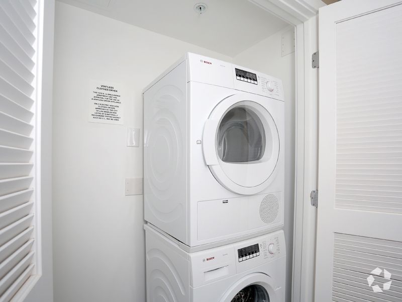 White stacked washer and dryer in a small, white laundry room. Door and blinds visible.