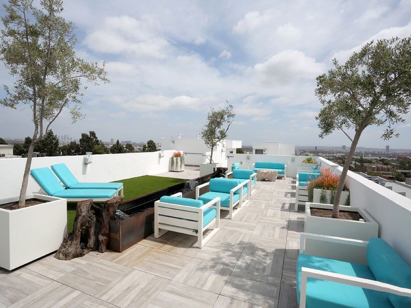 Rooftop patio with white furniture, teal cushions, trees, and city view under a cloudy sky.