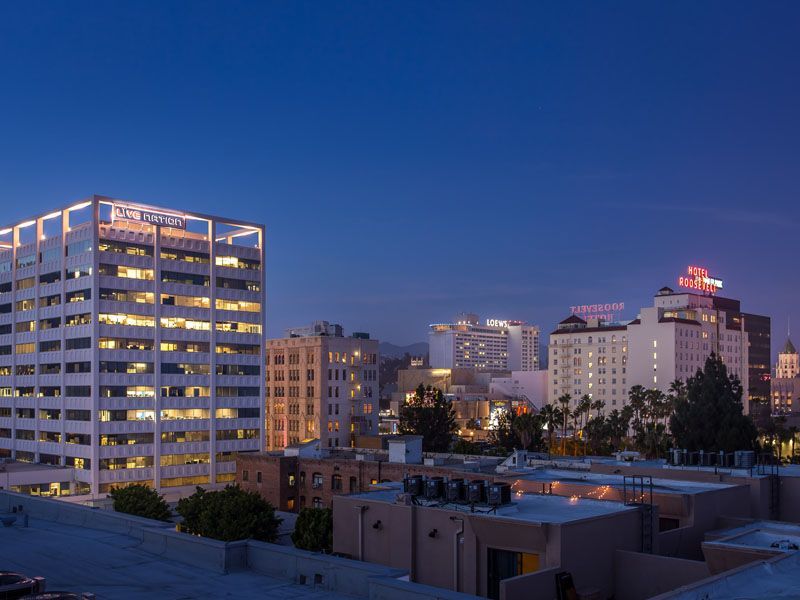 City skyline at dusk, with lit office buildings and a hotel sign against a darkening blue sky.