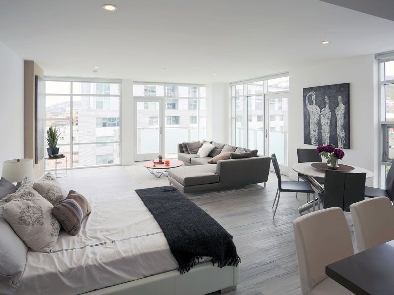 Modern open-concept living space with bed, couch, dining table, and large windows. Gray and white color scheme.