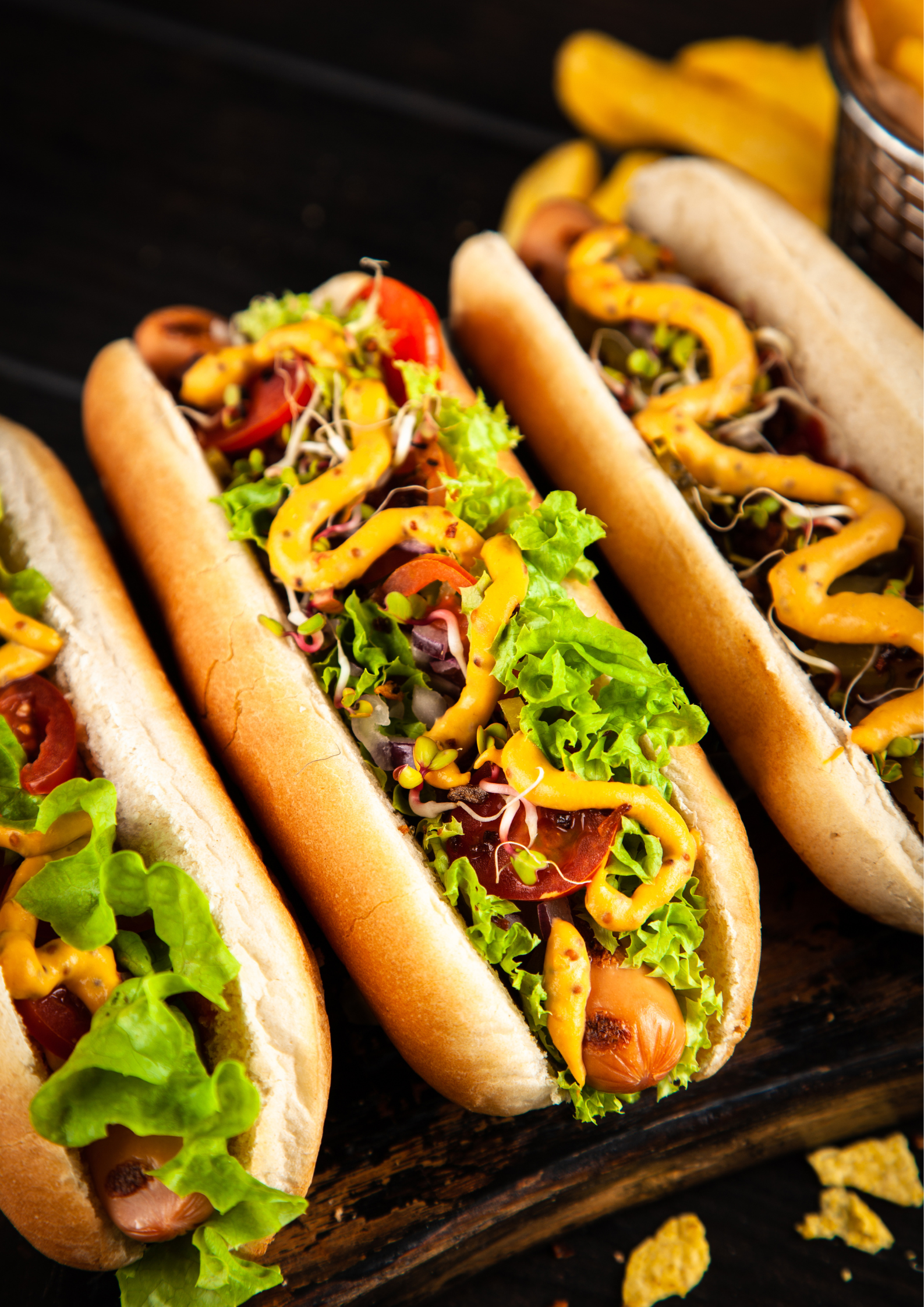 Three hot dogs with lettuce , cheese and tomatoes on a wooden table.