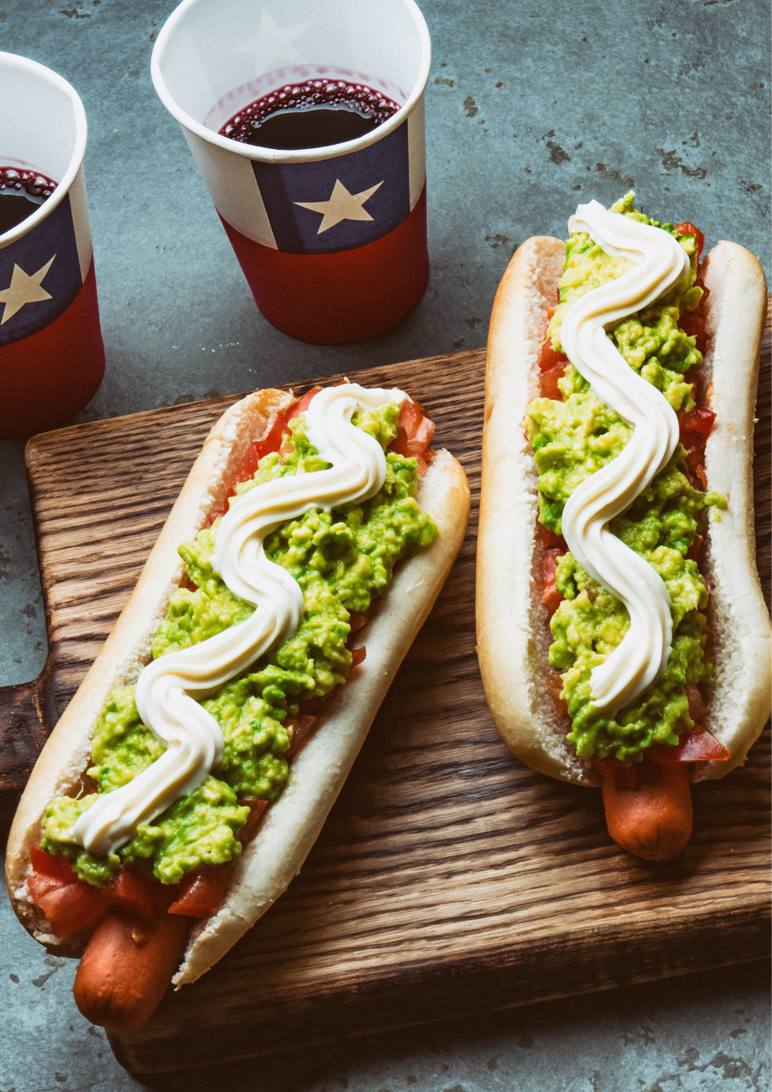 Two hot dogs with guacamole and mayonnaise on a wooden cutting board.