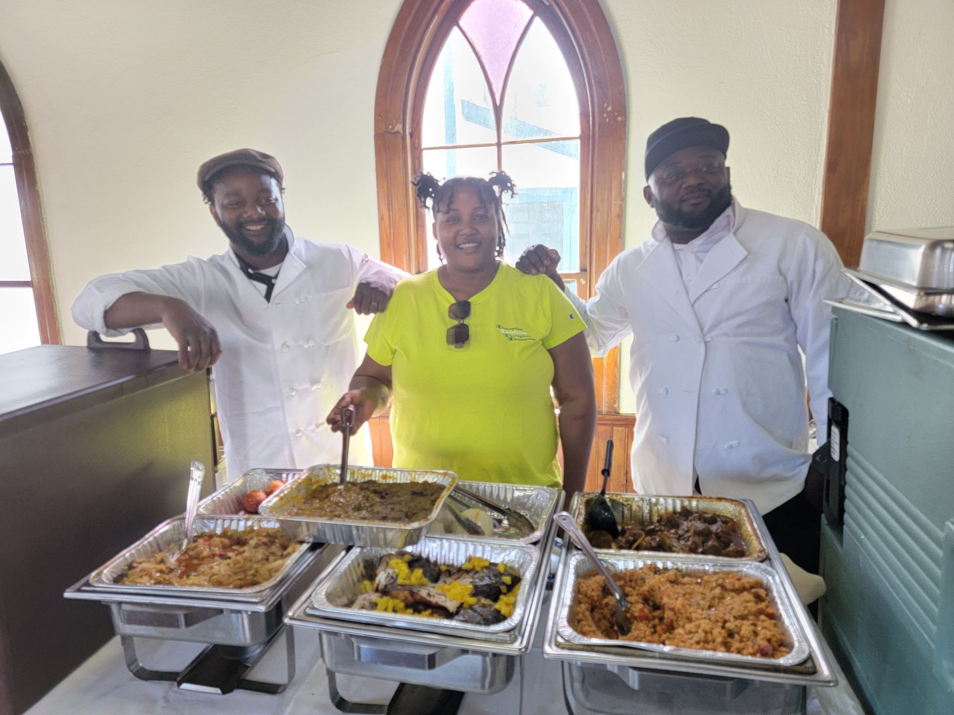 Two men and a woman are standing in front of a table with trays of food.