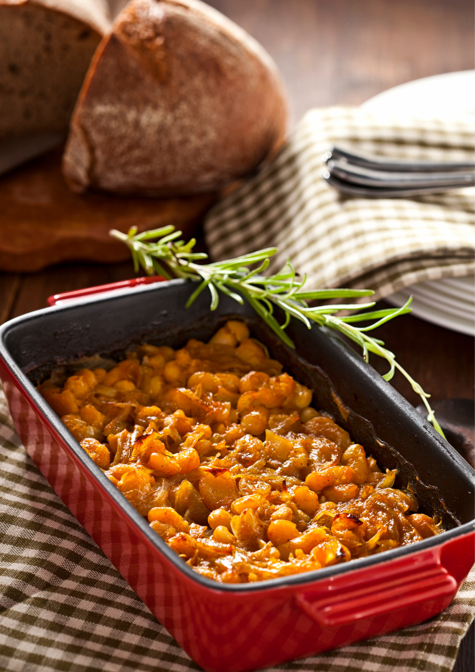 A red casserole dish filled with beans is on a table next to a loaf of bread.