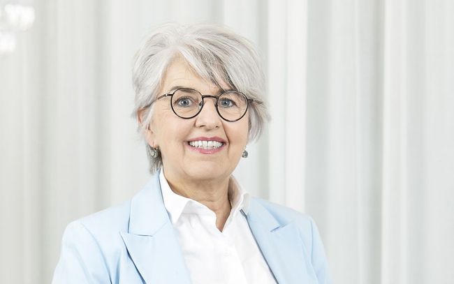 Une femme portant des lunettes et une veste bleue sourit à la caméra.