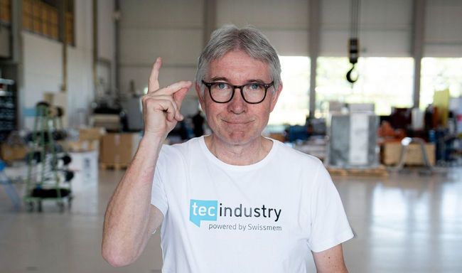 Un homme portant des lunettes et un t-shirt blanc pointe du doigt une usine.