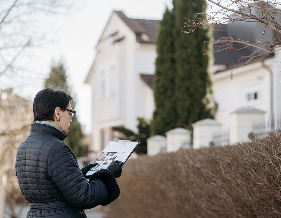 Person holding a clipboard beside a hedge in front of suburban houses