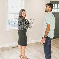 Two people in a bright room, one holding a clipboard and talking to the other near a window.