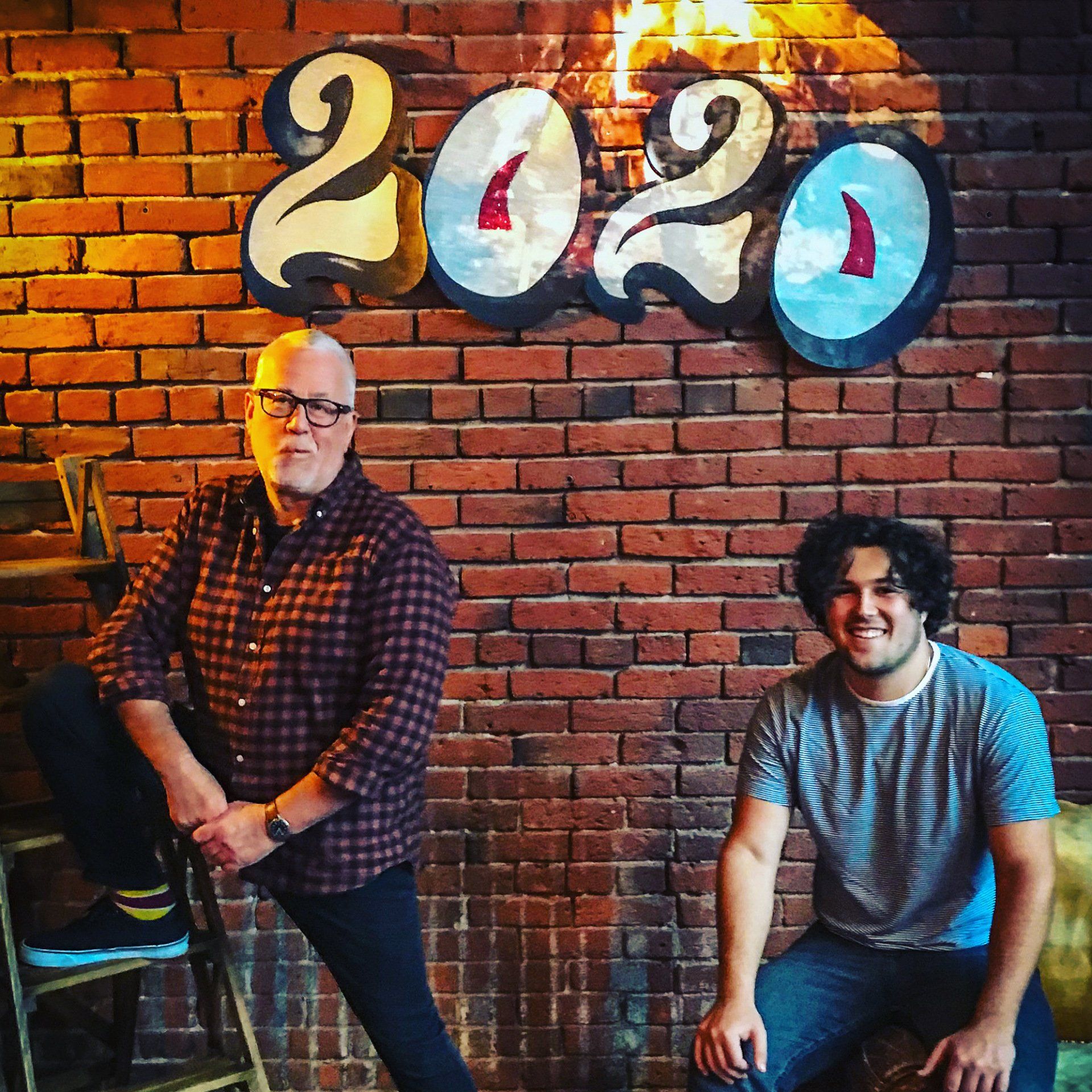 Two men pose by a brick wall with 