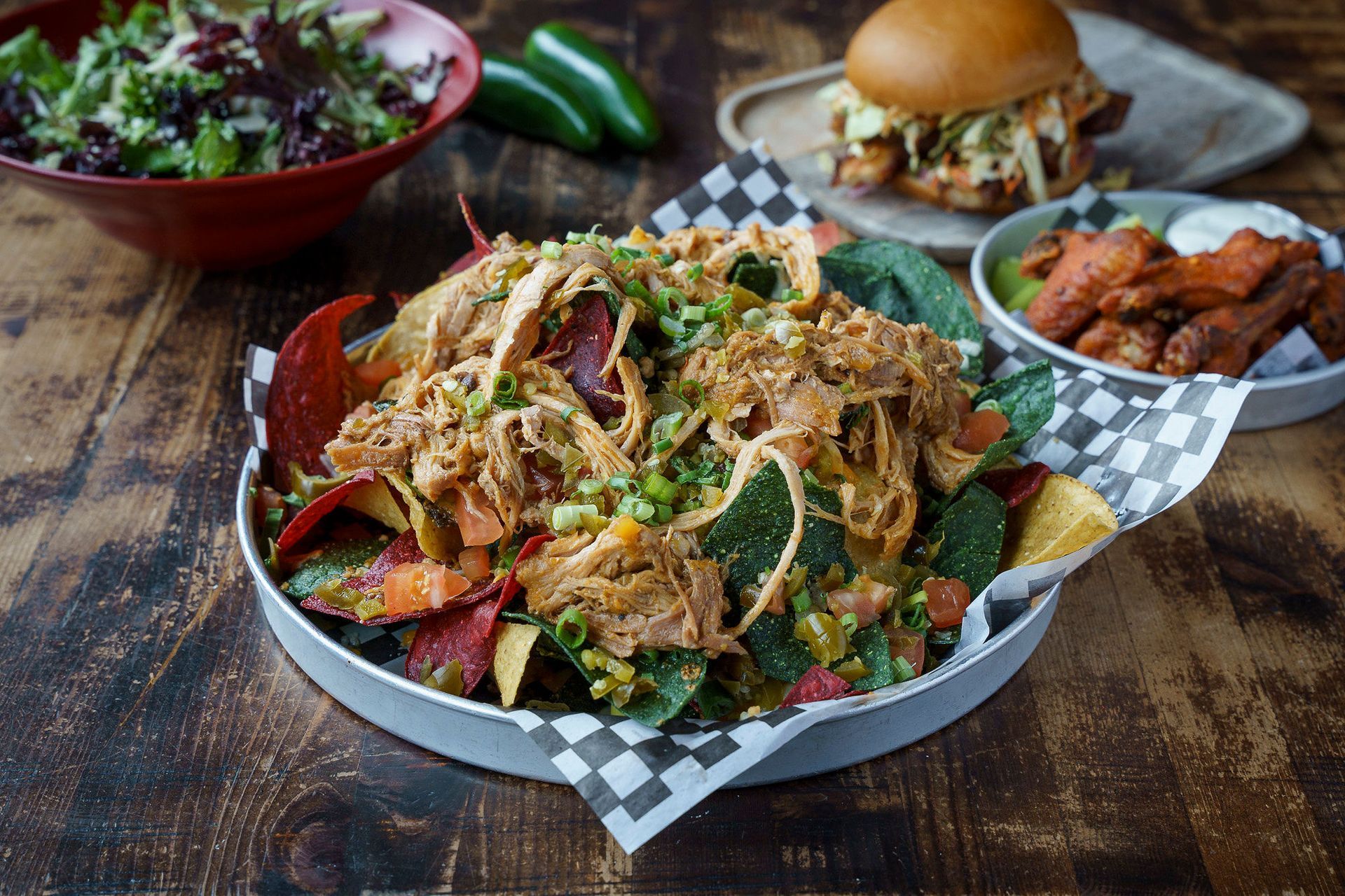 Nachos with pulled meat, salad, burger, and wings on a rustic wood table.
