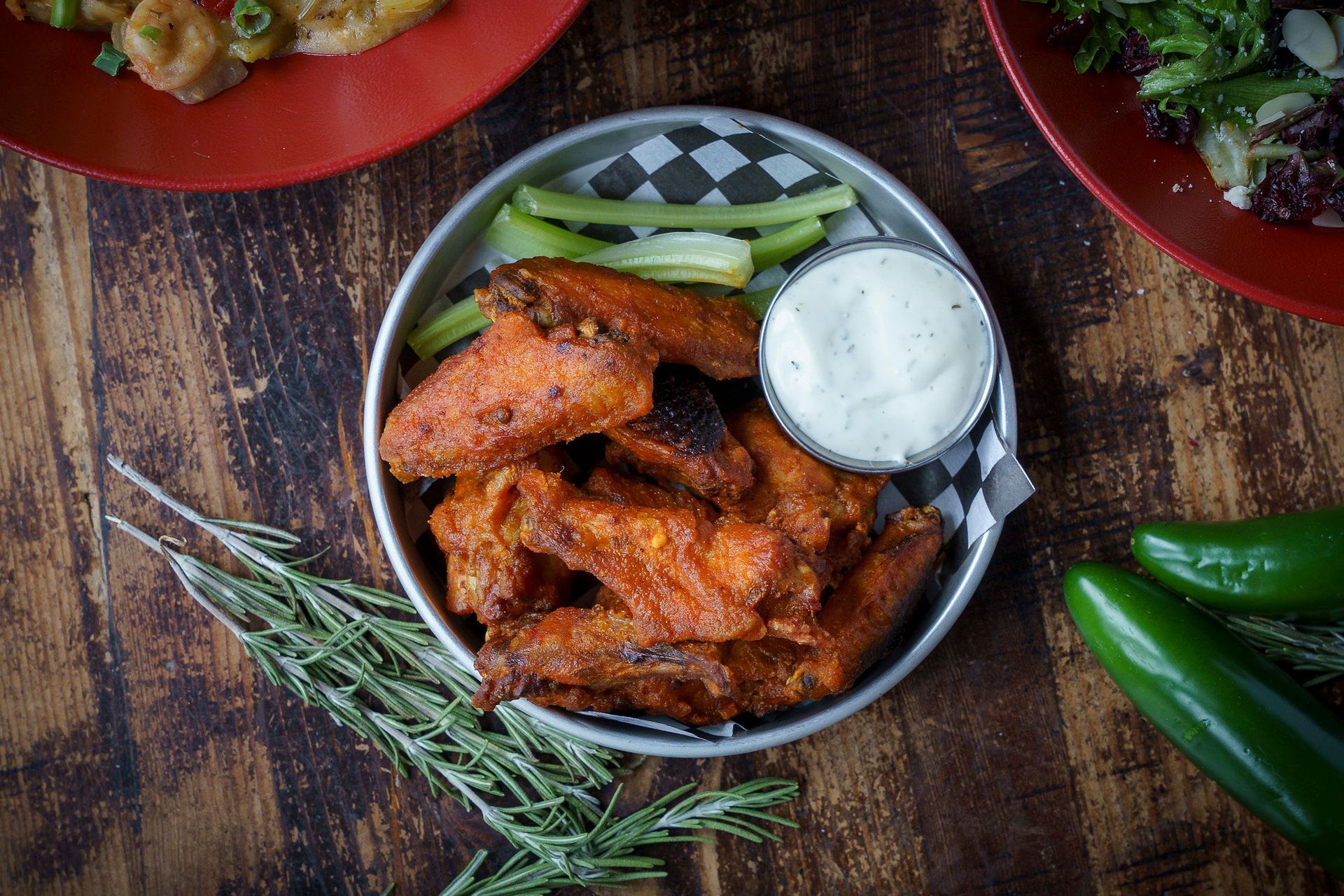 Chicken wings in metal bowl with ranch, celery, jalapenos on wood table.