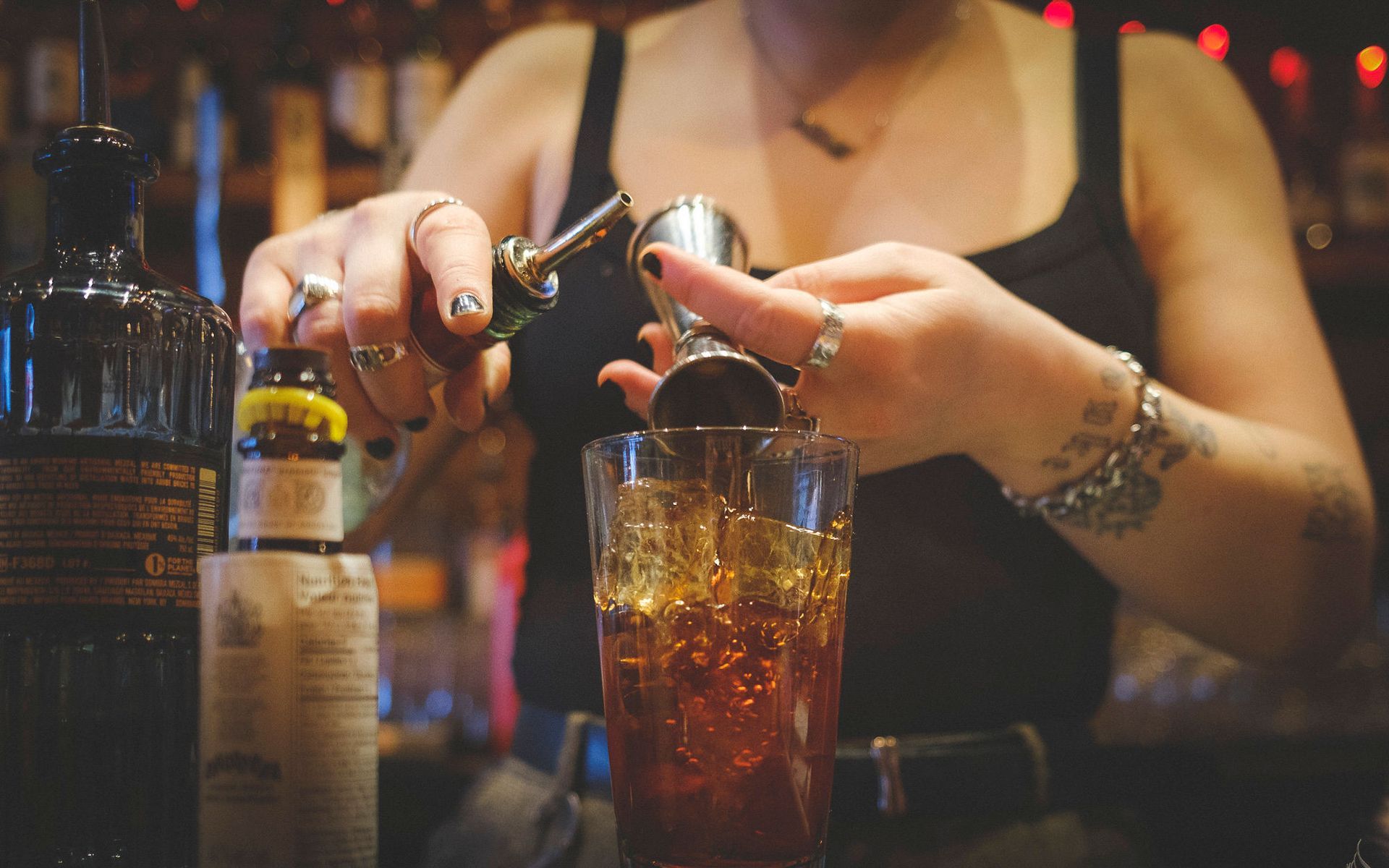 Bartender pouring drink from jigger into glass at a bar, wearing a black tank top with multiple rings and tattoos.