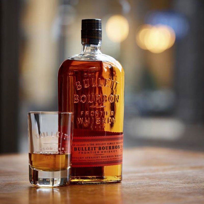 Bottle and shot glass of Bulleit Bourbon on a wooden surface. Amber liquid, dark bottle with red label.