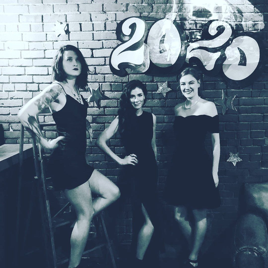 Three women in black dresses pose in front of a brick wall with 