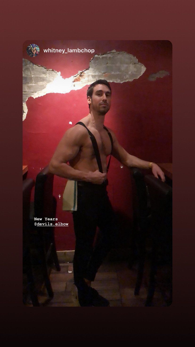 Shirtless man, suspenders, standing by chairs, red wall, dimly lit.