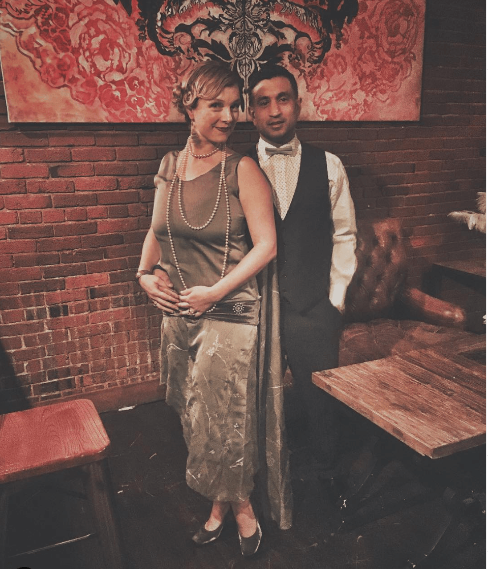 Couple in 1920s attire pose in front of a brick wall and art. She holds her stomach. He wears a vest and bow tie.