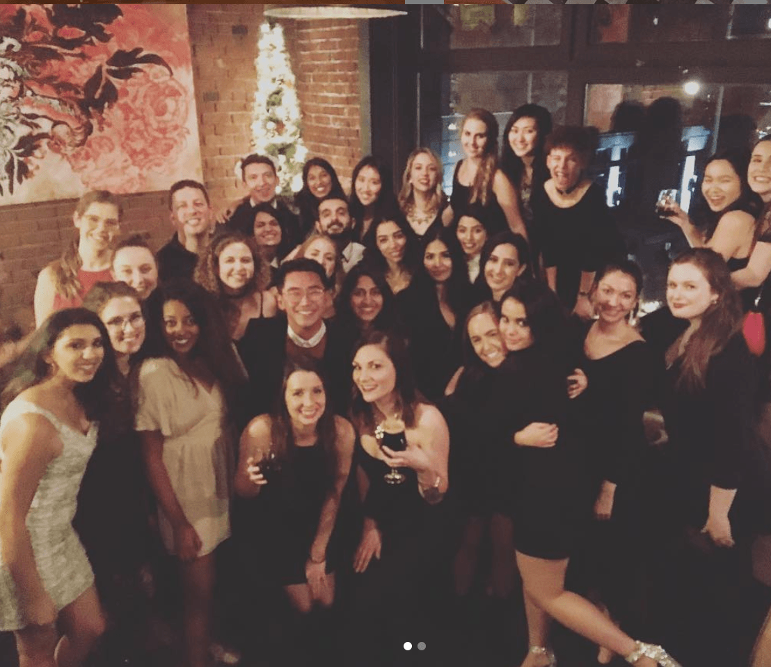 Group photo of diverse people at a party. Smiling and posing, some holding drinks, against a brick wall background.