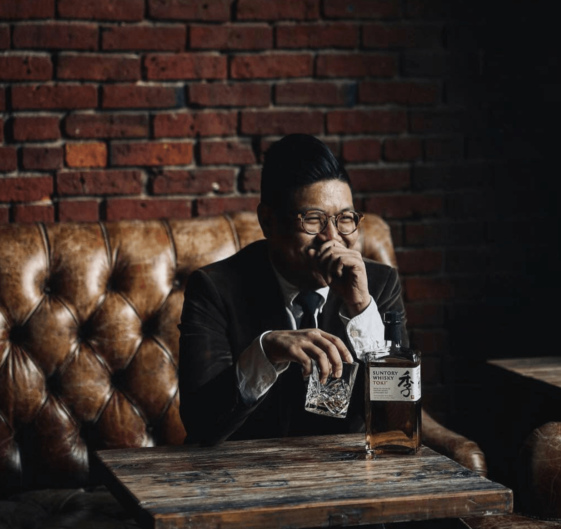 Man in suit with glasses, laughing while sitting at a table with whiskey, in front of a brick wall.
