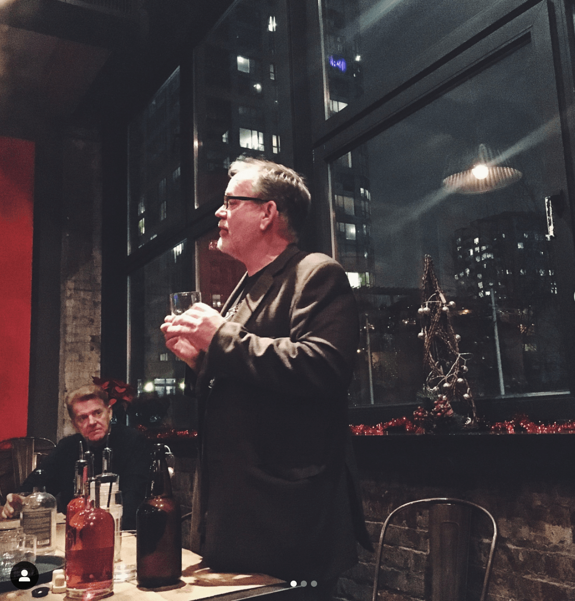 Man giving a speech indoors at night, holding a glass, with city lights visible through a window.