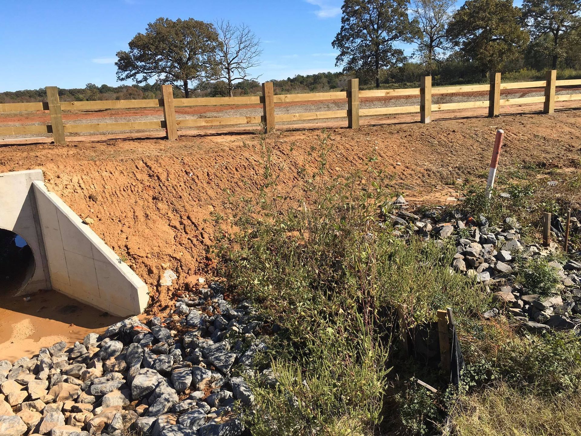 Dirt embankment with rocky drainage culvert beside a wooden fence in a rural field