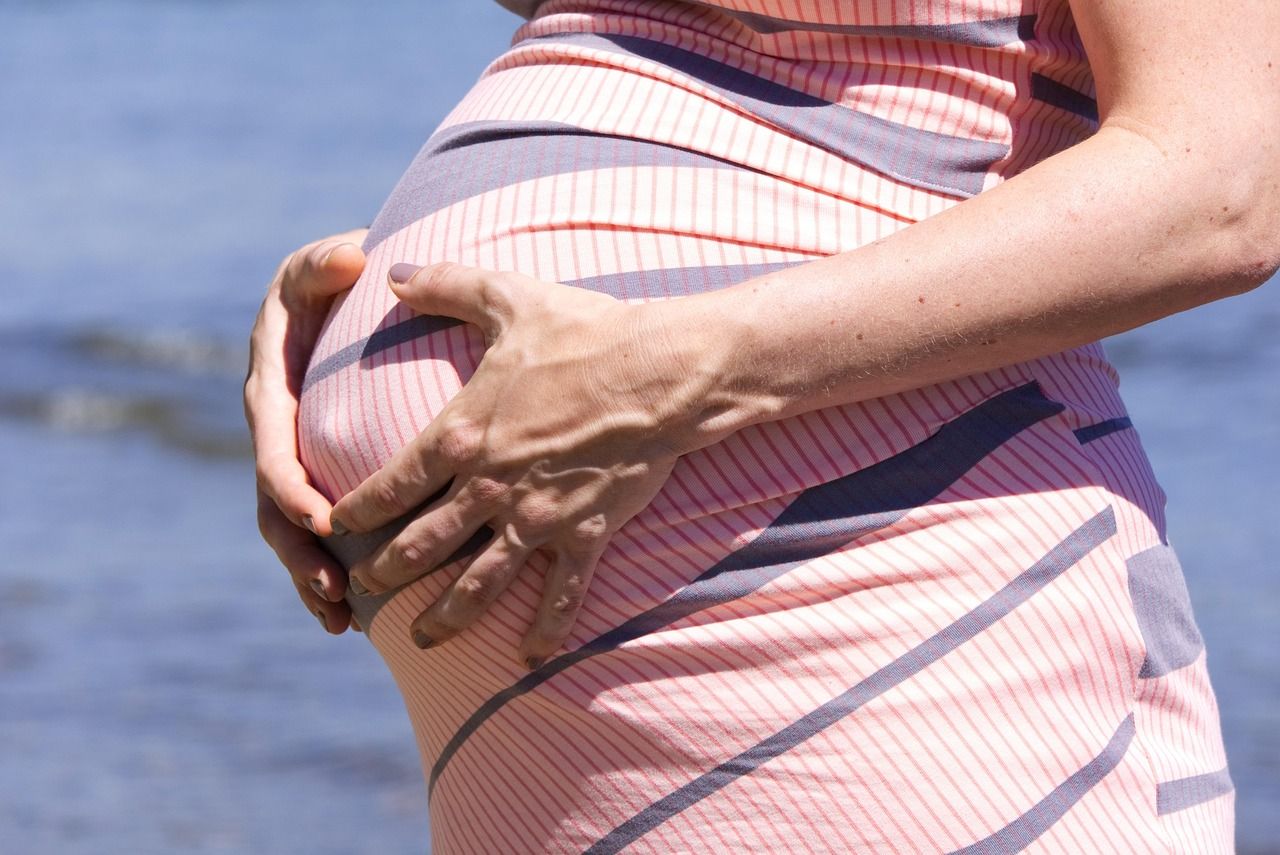 Pregnant woman in a pink dress, hands cradling her baby bump, by the water.