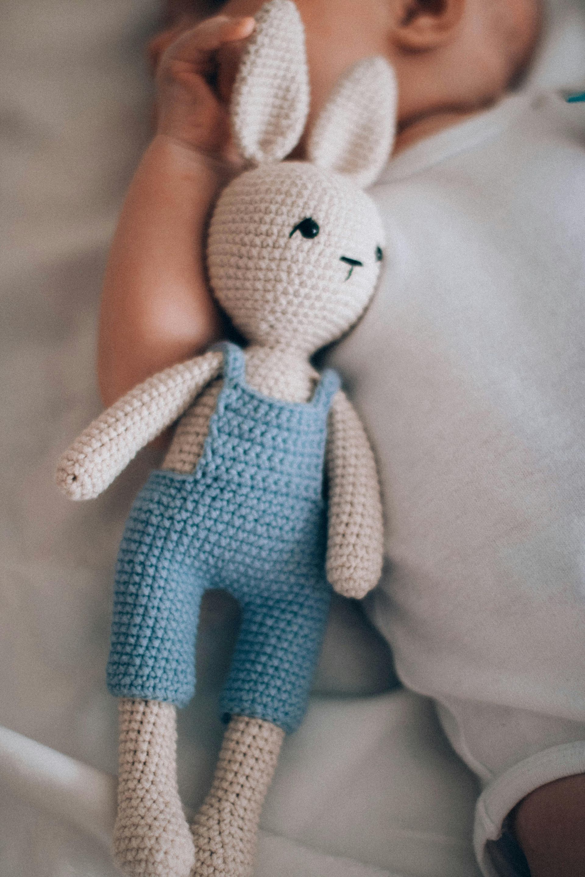 Crocheted bunny doll in blue overalls next to a baby in white onesie.