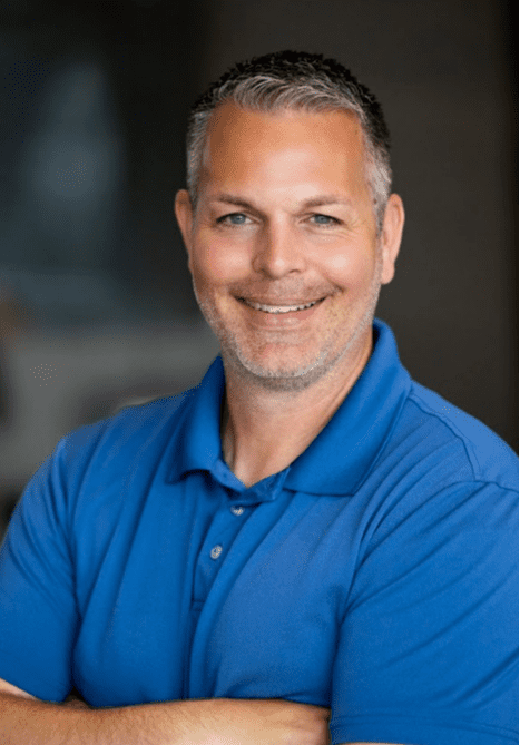 A man in a blue shirt is smiling with his arms crossed.