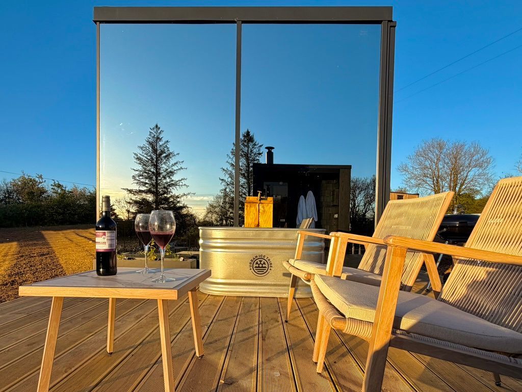 Wooden deck with chairs, table, wine glasses, and a fire pit under a clear blue sky at sunset